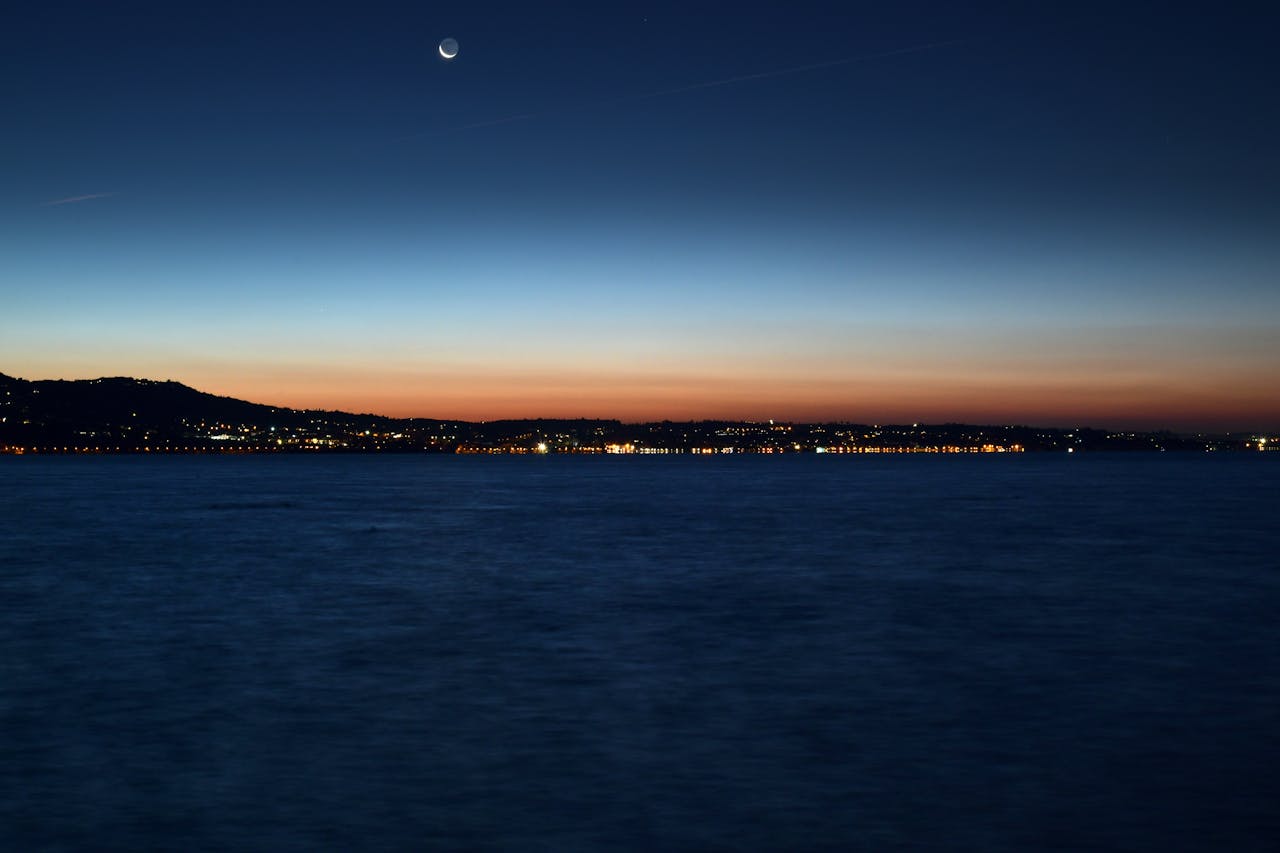 A tranquil view of Garda Lake under a crescent moon at twilight, capturing serene water and distant city lights.